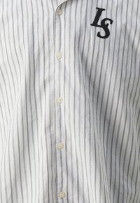 Camisa de botones en blanco con rayas verticales grises, con un bolsillo bordado con "LS" en negro, fabricada con tejido ligero.