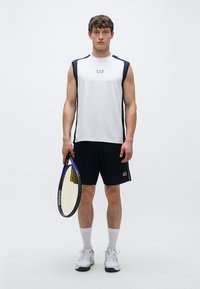 Hvit ermeløs treningsskjorte med svarte detaljer, kombinert med svarte shorts. Modellen holder en tenniskølle, iført hvite sokker og joggesko.