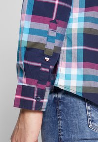Chemise à manches courtes à carreaux multicolores dans des tons de bleu marine, teal et bordeaux, avec un système de fermeture à boutons et un détail de bouton distinctif de Mickey Mouse.
