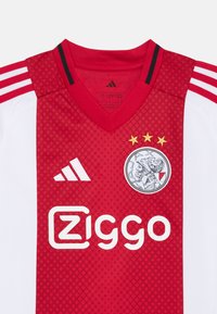 Maglietta da calcio rossa con maniche bianche, colletto a V, tre strisce nere sulle spalle, logo ricamato e testo di sponsorizzazione.