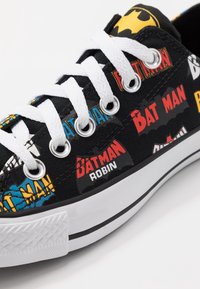 Schwarze Leinensneaker mit weißen Schnürsenkeln, die im Cartoon-Stil gestaltete Grafiken der Logos von "Batman" und "Robin" in Rot, Blau und Gelb zeigen.