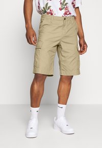 Beige Cargo-Shorts mit zwei Seitentaschen, einer lockeren Passform und einem Saum, der knapp über dem Knie endet. Getragen mit weißen Sneakers und Socken.