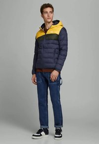 Casaco puff no azul marinho, amarelo e verde. Possui capuz, fecho com zip e bolsos laterais. Usado com jeans azuis e ténis pretos.