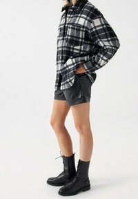 Person klädd i svart och vit rutig skjorta, svarta lädershorts och svarta snörkängor, står med en hand i fickan.