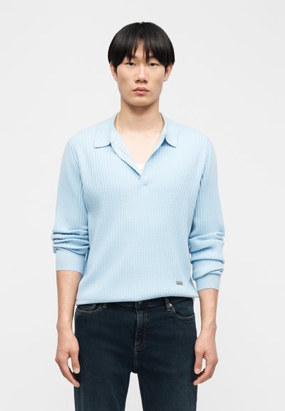 Jeune homme aux cheveux noirs et raides, portant un polo à manches longues bleu clair côtelé et un jean foncé, debout devant un fond clair uni.