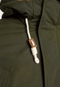 Olivgrön jacka med huva, som har ett strukturerat tyg, vit dragsko och beige trädetalj för justering. Tydliga sömmar synliga.