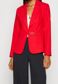 Rød skræddersyet blazer med en struktureret pasform, enkeltknap-lukning foran og guld kæde-detalje i taljen. Glat stof, klaffekrave.