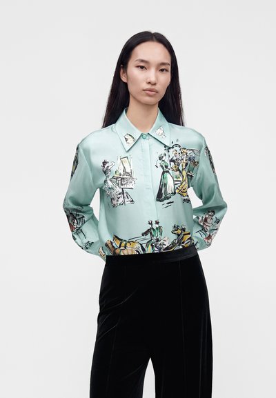 Femme aux longs cheveux foncés portant un chemisier bleu clair avec des illustrations au style vintage et un pantalon en velours noir, debout devant un fond uni.