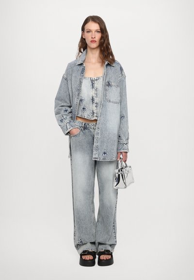 Šviesiai mėlynas džinsinis švarkas ir kelnių komplektas su gėlių siuvinėjimu. Sidabrinis metalinis rankinė, storos juodos šlepetės ir trumpa palaidinė. Tekstūruota medžiaga.