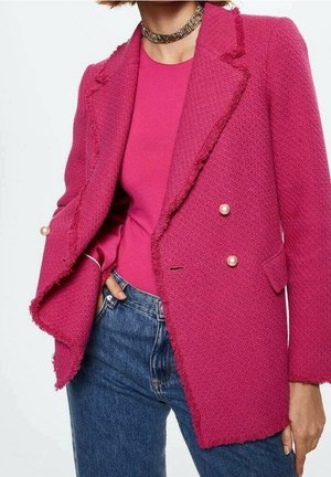Kvinde iført en struktureret fuchsia blazer med perleknapper, matchende fuchsia top, blå jeans og en gylden choker halskæde.