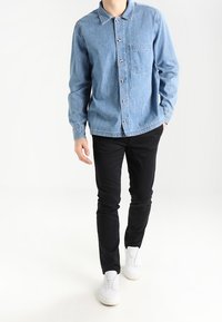 Světle modrá denimová košile s knoflíkovou přední částí, dlouhými rukávy a kapsou na prsou, v kombinaci s černými kalhotami a bílými teniskami.