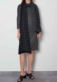 Femme portant une robe gris foncé longueur genou, des mules à talons noires à bout ouvert et une longue écharpe noire et grise à motif.