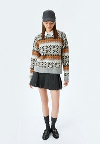 Strickenes Sweater mit einem gemusterten Design in Creme, Schwarz und Rost. Kombiniert mit einem grauen Faltenrock, grauen Socken und schwarzen Loafers.