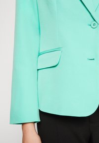 Turkoss blazer med enkel knapp, fickor med klaff och lång ärm, bärs över svarta byxor.
