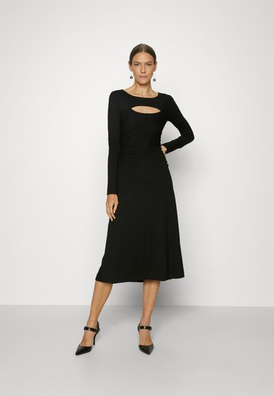 Μαύρο μακρυμάνικο φόρεμα με μήκος μέχρι τα μισά της γάμπας, με μπροστινές κοπές και εφαρμοστή σιλουέτα, φτιαγμένο από μαλακό ύφασμα.