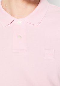 Polo rose clair avec un col côtelé, une patte à deux boutons et un logo subtilement embossé sur le côté gauche de la poitrine.