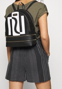 Black quilted backpack with white logo detail, gold hardware, and textured bottom. Worn with a gray shirt and patterned shorts.
