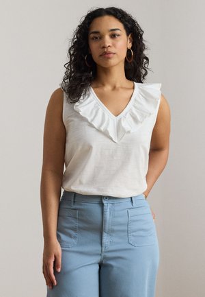 Femeie cu păr creț și întunecat, purtând cercei aurii hoop, top alb fără mâneci cu decolteu în V și volane, și pantaloni bleu deschis cu talie înaltă.