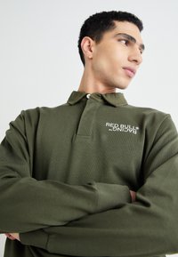 Zelena bombažna polo majica z gumbnim ovratnikom, na katerem je bel logotip na prsih. Dolgi rokavi z rebrastimi manšeti. Gladka tekstura.