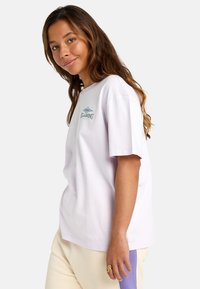 Svetlofialové bavlnené tričko so malým grafickým logom na hrudi a voľným strihom, skombinované so smotanovými nohavicami s fialovými akcentmi.