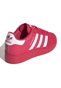 Röd Adidas Superstar-sneaker med vita ränder och logotyp på hälen, sedd från ett bakåtlutat perspektiv mot en vit bakgrund.