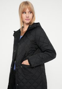 Czarna pikowana kurtka z kapturem, z zapięciem na zamek błyskawiczny, zatrzaskami i kieszeniami po bokach. Wykonana z gładkiego, izolowanego materiału.