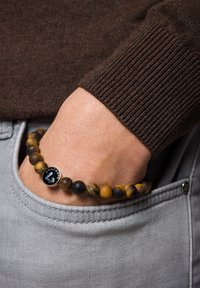 Bransoletka z koralików wykonana z kamieni tygrysiego oka w różnych odcieniach brązu i czerni, z metalowym zawieszką z logo.