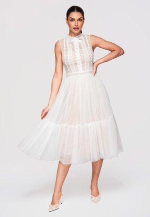 Frau trägt ein ärmelloses weißes Spitzen-Midikleid mit Schleifen-Halsausschnitt, transparentem gepunktetem Rock und weißen High Heels, posiert mit der Hand in der Hüfte.