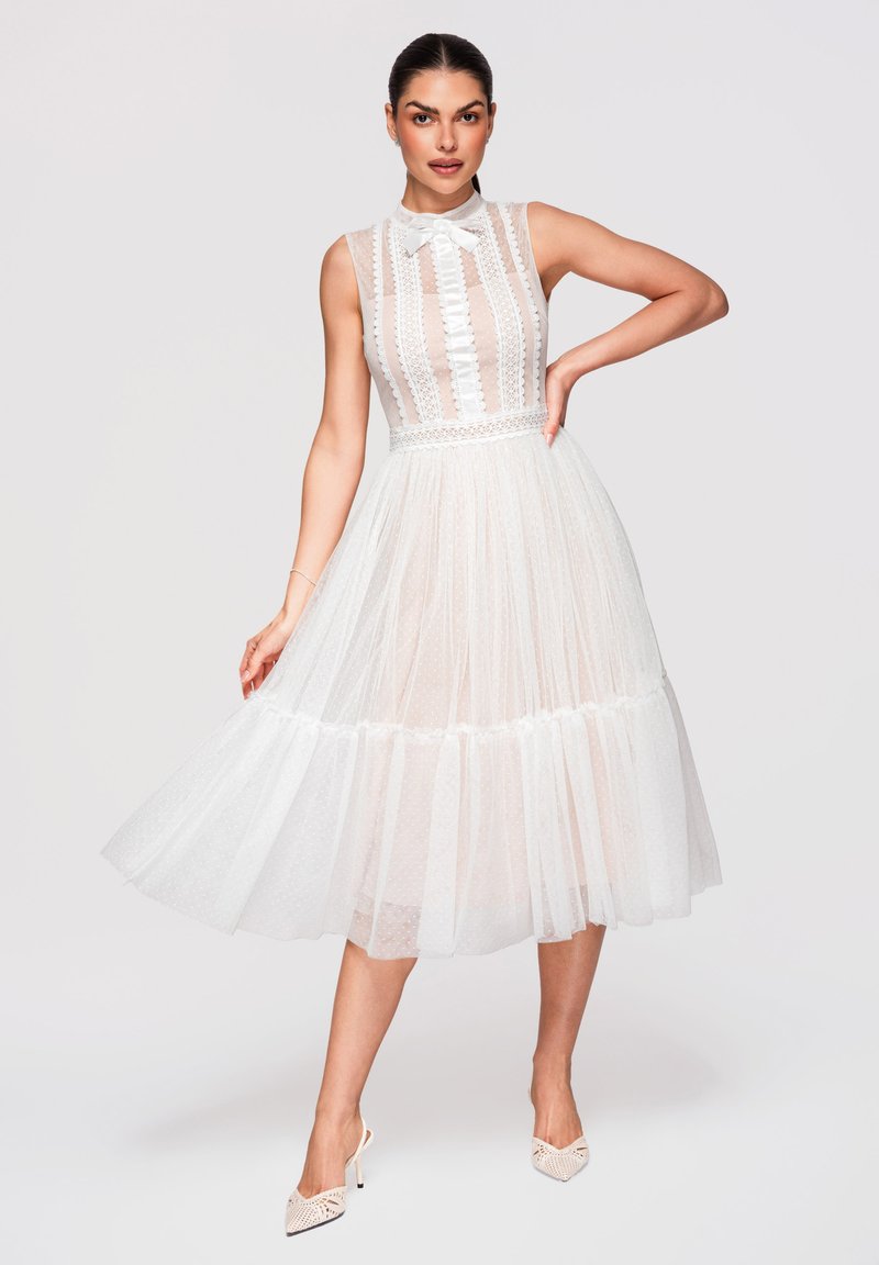 Femme portant une robe midi blanche en dentelle sans manches avec un détail nœud au col, une jupe transparente à pois et des chaussures blanches à talons hauts, posant la main sur la hanche.