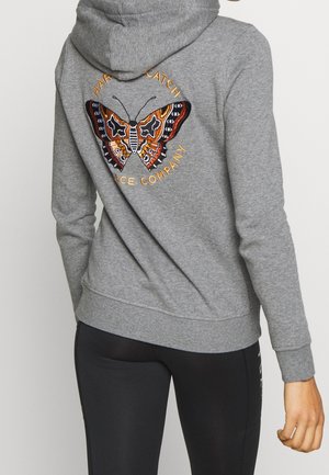 Personne portant un sweat à capuche gris avec un papillon coloré et le texte "Hard to Catch Dance Company" brodé dans le dos.