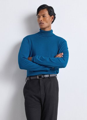 Uomo con dolcevita blu e pantaloni neri con cintura nera, in piedi con le braccia incrociate, guarda a sinistra su uno sfondo bianco semplice.