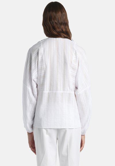 Femme aux cheveux bruns ondulés portant un chemisier blanc ample à manches longues texturé et un pantalon blanc, se tenant de dos sur un fond uni.
