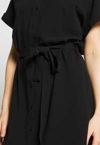 Black button-down dress featuring short sleeves, a belted waist, and textured fabric. Buttons are black, and the belt is tied at the front.