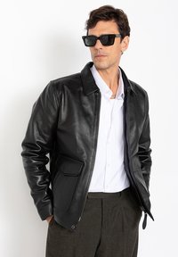 Veste en cuir noir avec col, fermeture éclair frontale et poche latérale. Portée sur une chemise blanche, associée à un pantalon foncé et des lunettes de soleil.