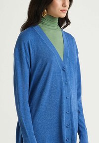 Cardigan bleu à col en V, boutonné sur le devant, avec des manches longues, confectionné dans un matériau texturé et doux, porté sur un col roulé vert.