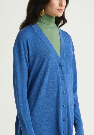 Cardigan bleu à col en V, boutonné sur le devant, avec des manches longues, confectionné dans un matériau texturé et doux, porté sur un col roulé vert.