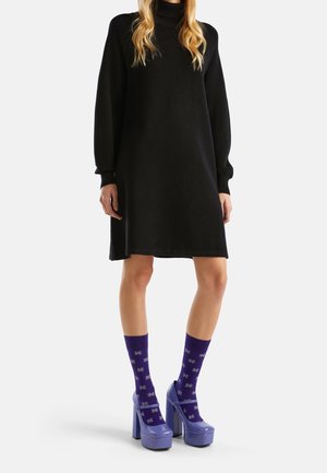 Kobieta ubrana w czarną sukienkę z długim rękawem i golfem, fioletowe skarpetki w wzory oraz fioletowe buty na platformie, stojąca na białym tle.