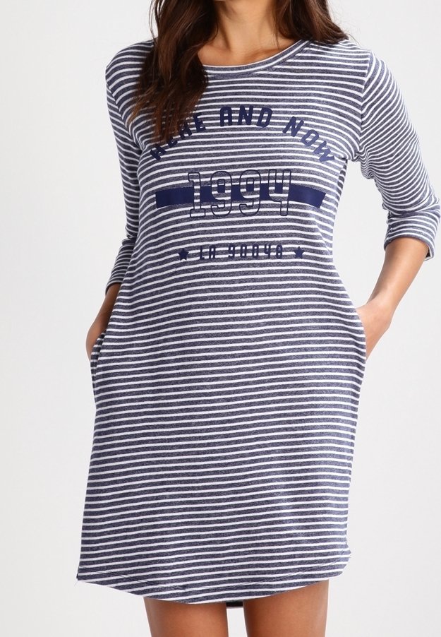 Femme portant une robe aux genoux à rayures bleu marine et blanches avec des manches 3/4 et l'inscription « THEN AND NOW 1994 » sur le devant, mains dans les poches.