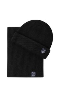 Melns ribots adījuma beanie un atbilstoša šalle ar nelielām taisnstūra formas zīmola etiķetēm šalles malā un beanie aprocē.
