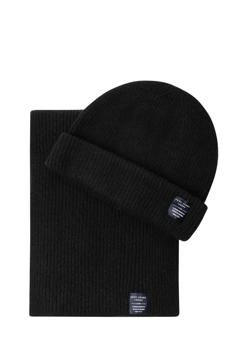 Melns ribots adījuma beanie un atbilstoša šalle ar nelielām taisnstūra formas zīmola etiķetēm šalles malā un beanie aprocē.