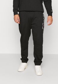 Zwarte sweatpants gemaakt van een katoenmengsel, met een taps toelopend ontwerp en witte logovoorzieningen langs de linkerkant. Draagt met witte sneakers.