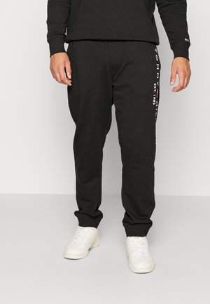 Pantalons de survêtement noirs en tissu mélangé coton, avec un design fuselé et des accents de logo blancs sur le côté gauche. Portés avec des baskets blanches.