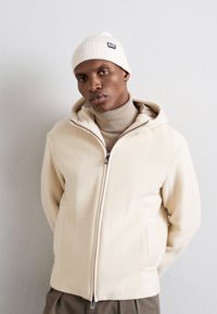 Beżowa bluza z kapturem na zamek błyskawiczny o teksturowym wykończeniu, noszona na jasnym golfie, w połączeniu z białą dzianinową czapką.
