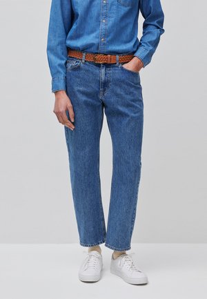 Person trägt blaue Jeans aus Denim und ein dazu passendes Hemd mit braunem geflochtenem Gürtel und weißen Sneakers, steht vor schlichtem Hintergrund.