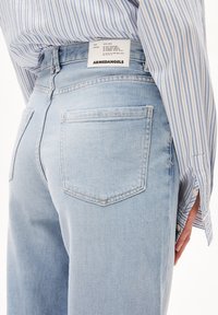 Lysblå denimbukser med baglommer og et hvidt "ARMEDANGELS" mærke, båret med en blå og hvidstribet skjorte.
