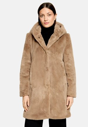 Femme portant un manteau en fausse fourrure beige avec capuche sur un col roulé noir, debout devant un fond clair uni.