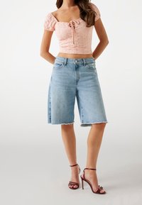 Kvinde iført lysblå knælange denim shorts, lyserød crop top med puffærmer i broderi og mørkerøde højhælede sandaler.