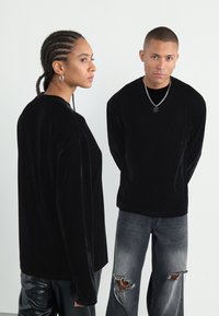 Cămăși lungi din catifea neagră purtate cu blugi gri uzati. Hainele au o textură netedă și o croială lejeră, având detalii de design minime.