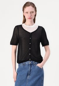 Svart lätt stickad cardigan med korta ärmar, v-ringning, knäppning och ribbad kant, bärs över en vit topp och matchas med denim.