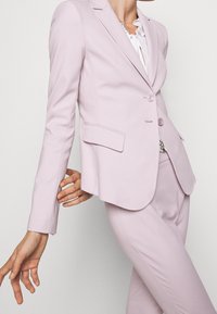 Vaaleanpunainen räätälöity bleiseri, jossa on yksi nappi, lovet kaulukset ja kaksi etutaskua, yhdistettynä samansävyisiin istuviin housuihin.